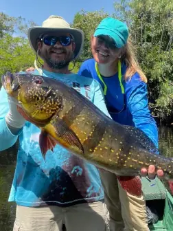 A couple holding a large peacock bass caught on a scenic river in Brazil. The image captures the thrilling beauty of fishing in Brazil.