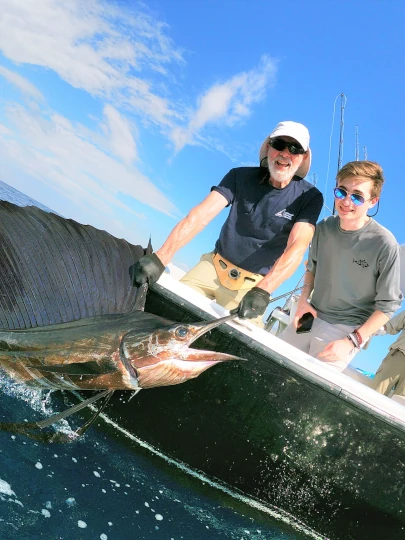 Two generations of anglers hold a large pacific sailfish alongside a boat in Guatemala.